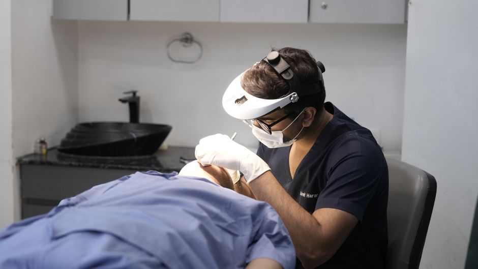 A dimmer treatment room with a dark basin, wall cabinets and a reclined chair, where a clinician in loupes works.