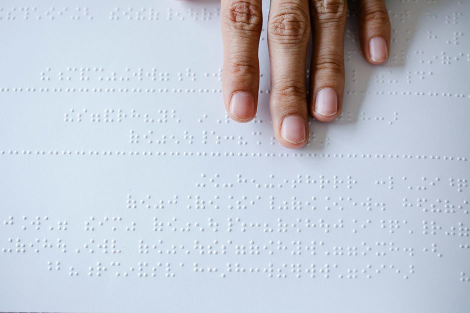 Four fingertips moving across a page of braille, photographed from directly above.