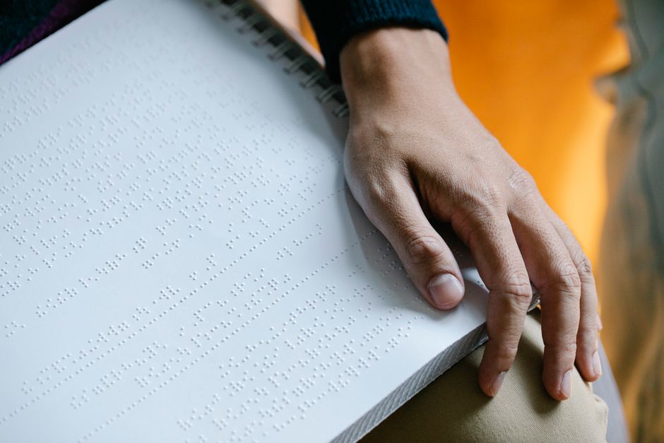 A person holding an open braille volume across their lap, reading with one hand.