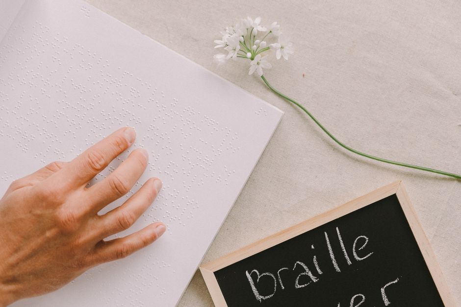 A hand flat on an open braille book beside a small chalkboard and a sprig of white flowers, on a linen cloth.