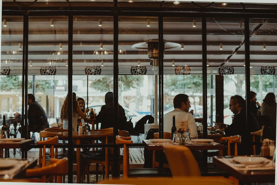 A restaurant interior seen through tall steel-framed glazing, tables and bentwood chairs in warm light.