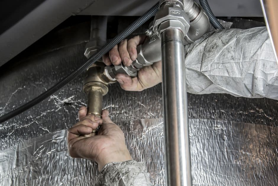 A plumber's hands tightening a chrome trap fitting on pipework beneath a sink.