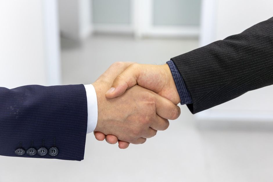 Two people shaking hands, photographed from the forearms down against a pale wall.