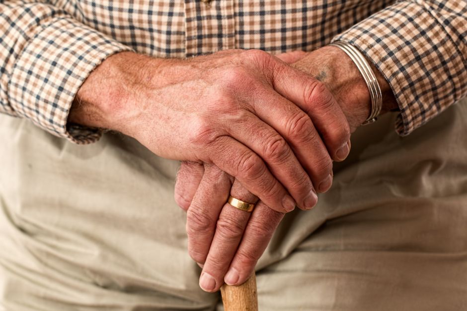 An older person's hands resting one over the other on the handle of a wooden walking cane.