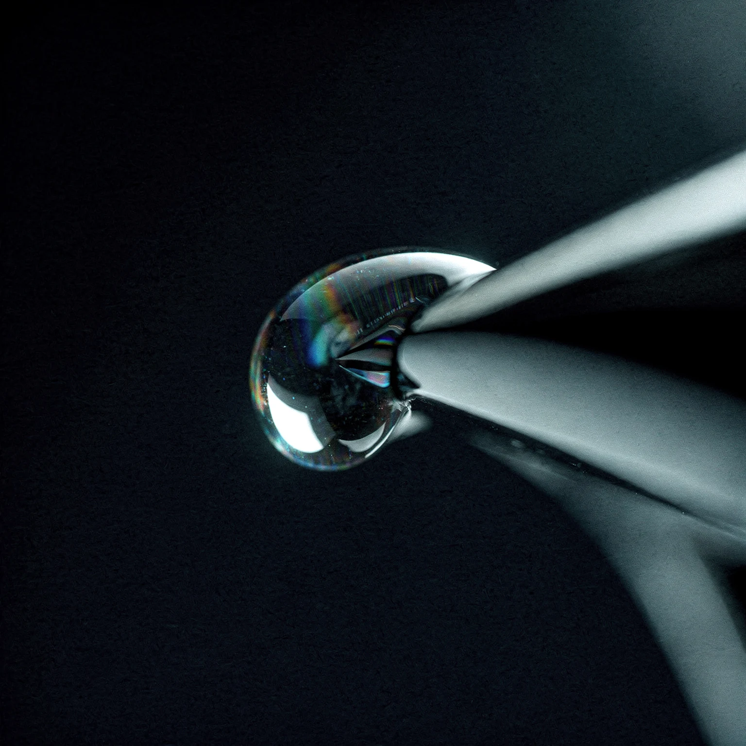 A single droplet at the edge of a chrome form, thin-film iridescence breaking across it.